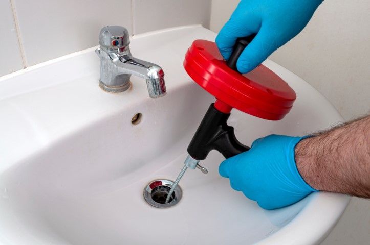 A person wearing blue gloves uses a red handheld plumbing snake to clear a clog in a white bathroom sink drain.