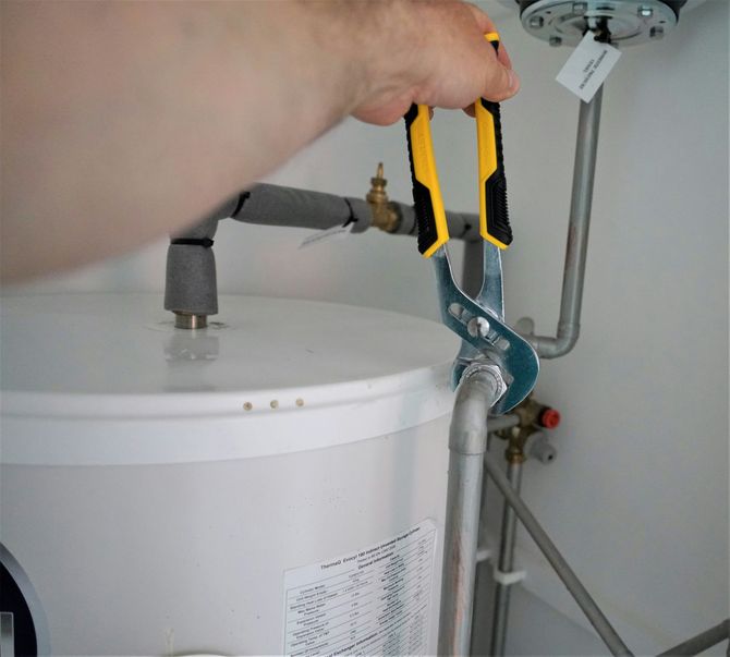 A hand holds yellow and black pliers, tightening a metal pipe fitting on top of a white water heater tank.