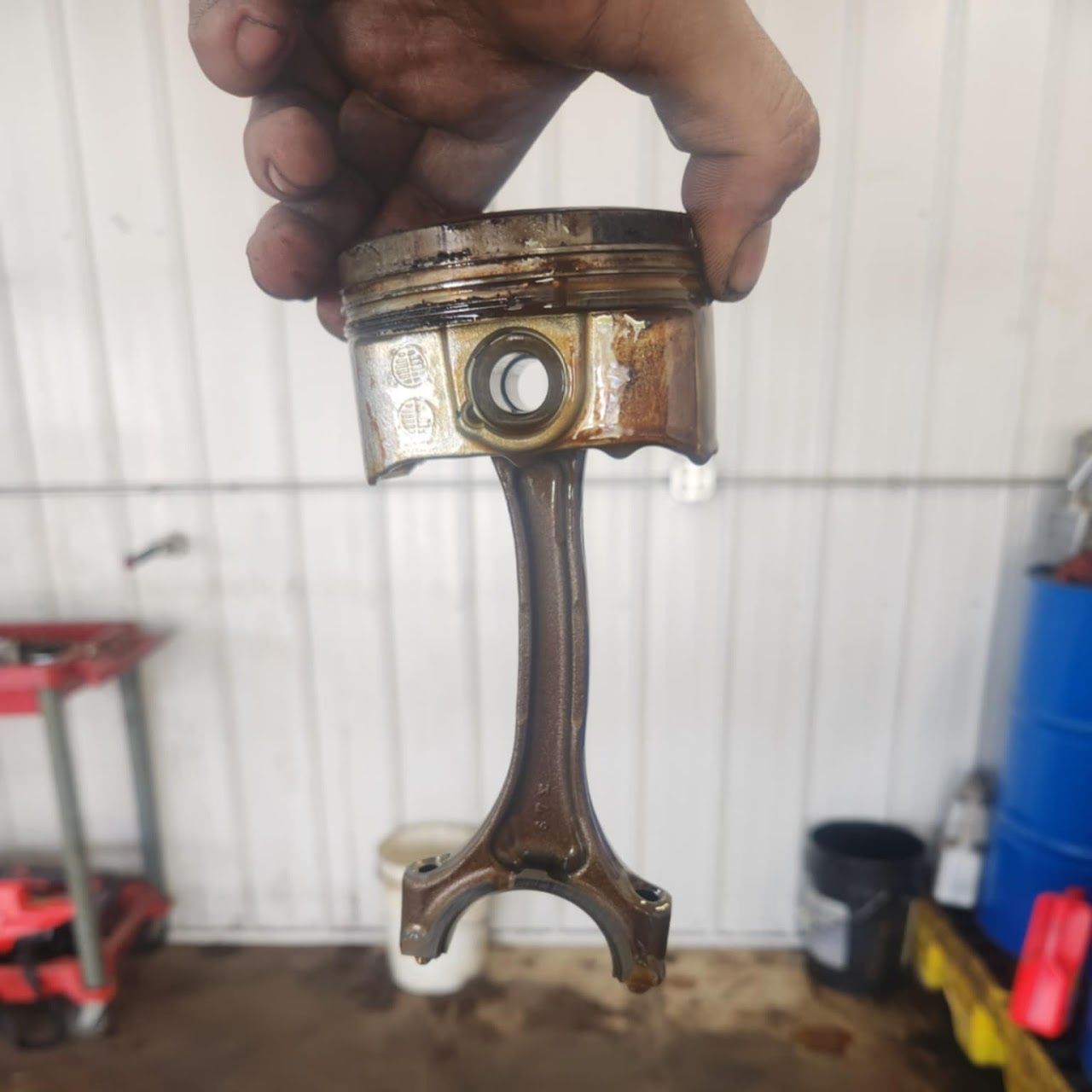 Damaged piston and connecting rod held up, oily, in a garage | Lucky Autolabs