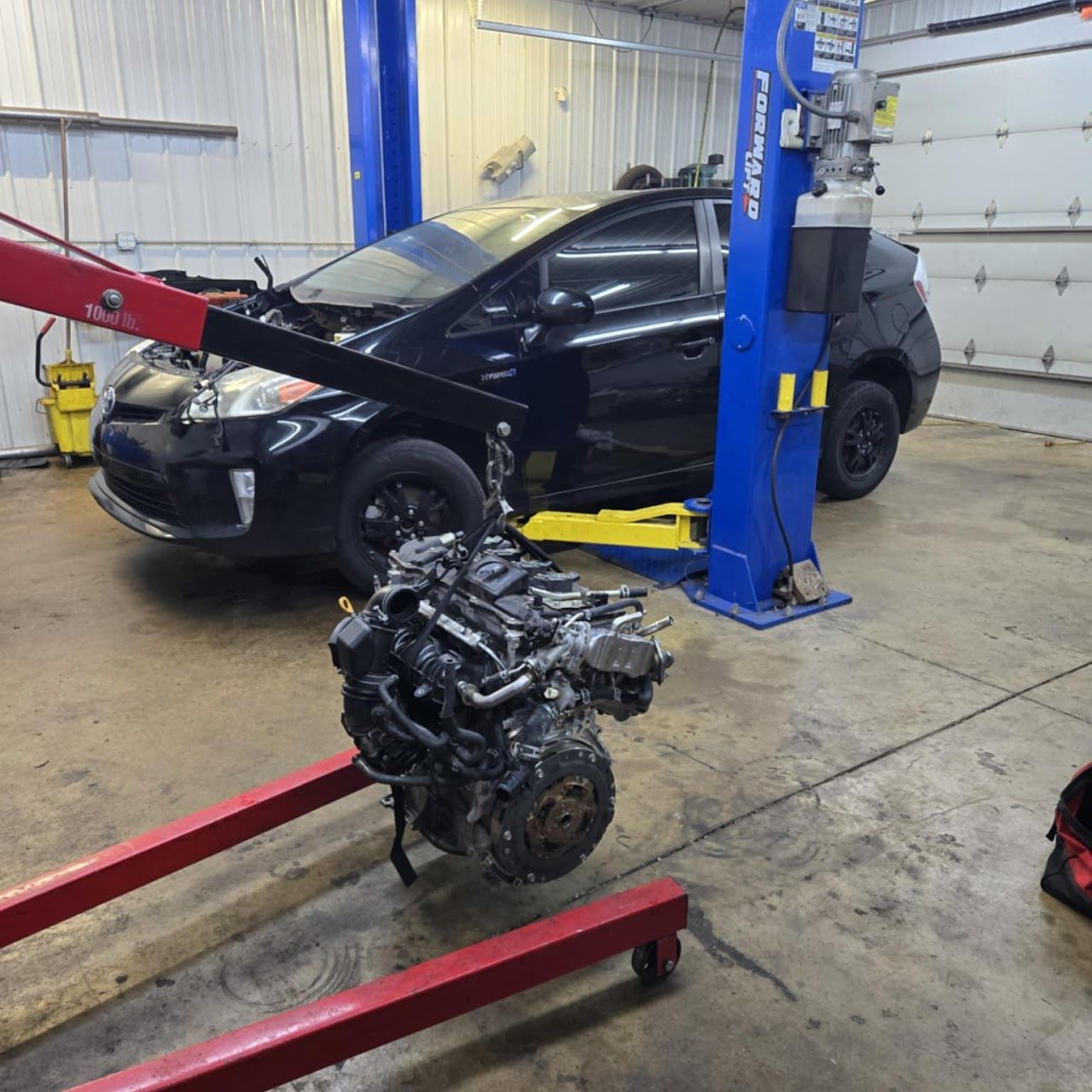 Black Prius with its engine removed in an auto repair shop. The engine hangs from a red hoist | Lucky Autolabs
