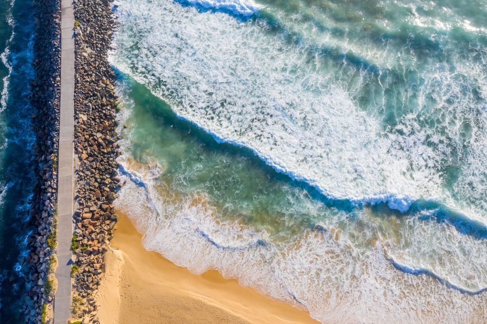 Ocean waves crashing on a sandy beach, next to a stone breakwater and a pathway. — Serene Therapy with Lana In Paddington, NSW