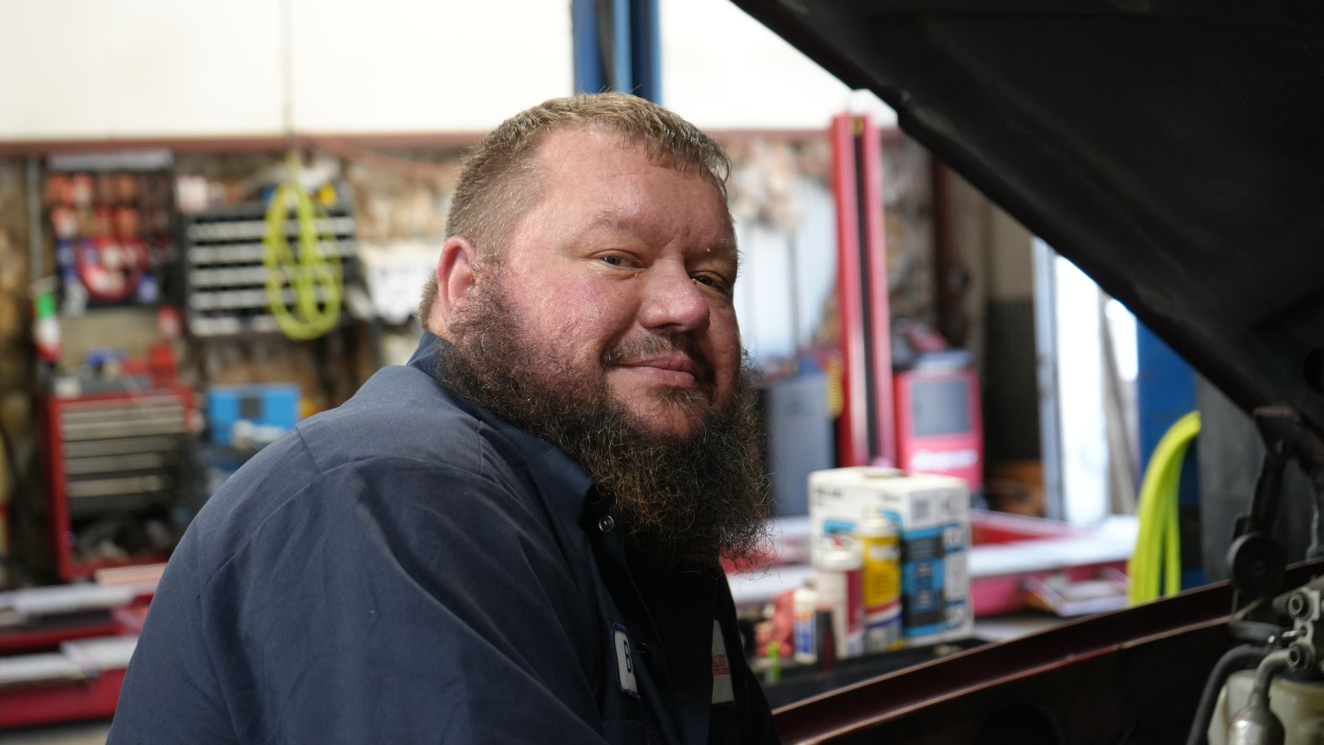A man with a beard is standing in front of a car with the hood up.