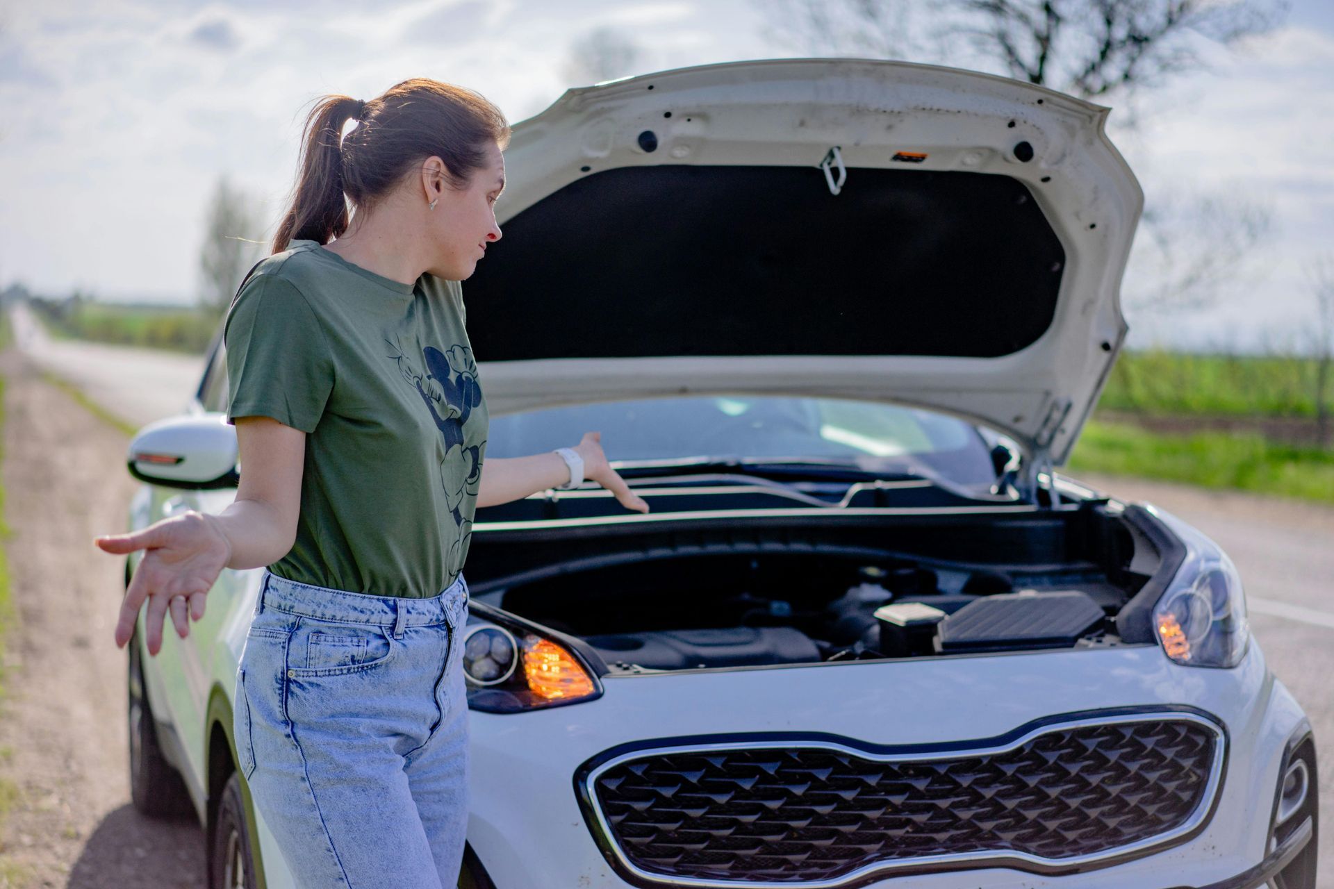 Woman looking frustrated in front of a white car with its hood up on a road.