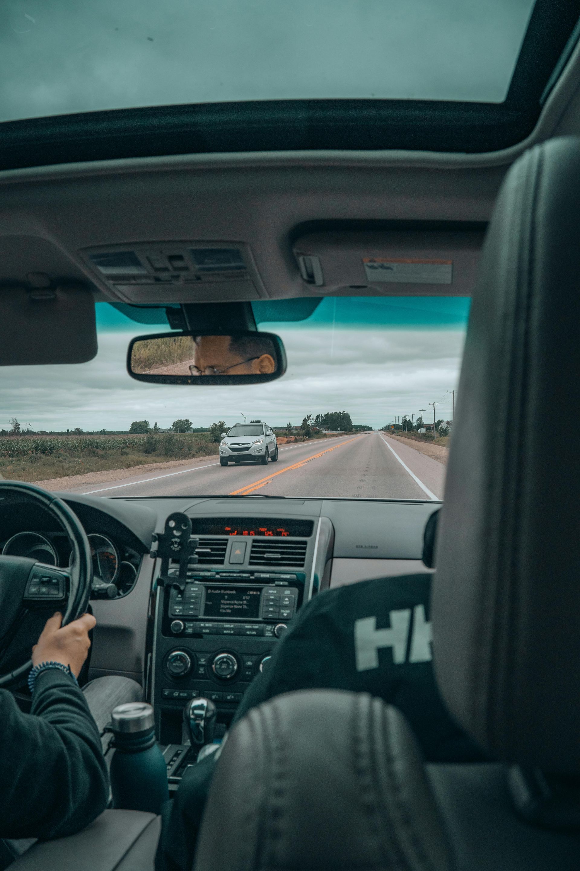 Interior car view, driving on a road. A car in the distance, blue sky, dashboard and steering wheel visible.