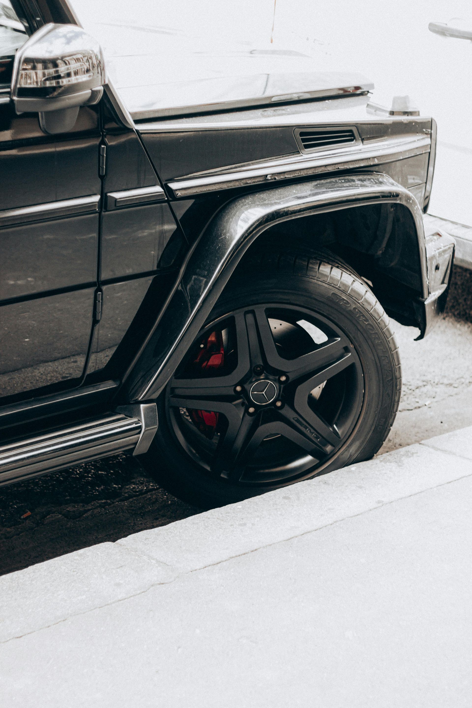 A close-up view of the front tire and fender of a matte black Mercedes-Benz G-Wagon parked next to a concrete curb.