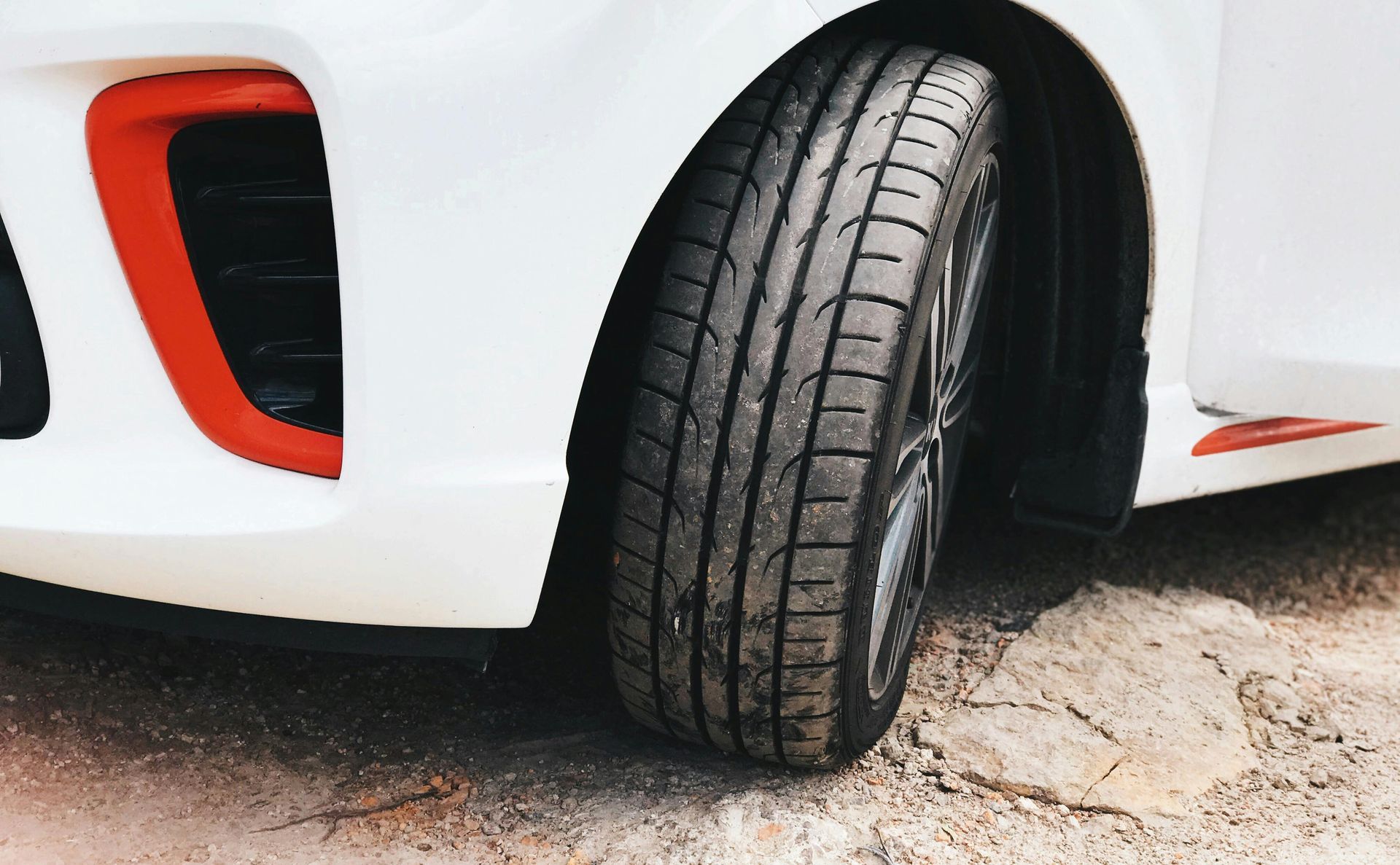 Close-up of a white car's front tire with black rims, parked on a dirt surface.