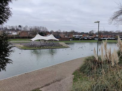Lake with an island and a white canopy structure. Ducks swim in the water, with townhouses in the background.