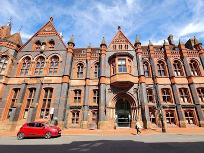 Ornate red-brick building with arched entrance, red car parked in front, person walking by.