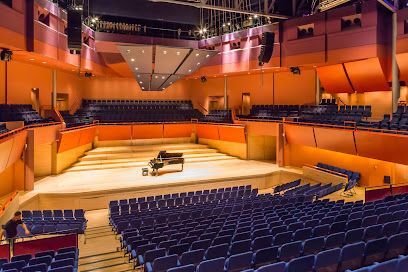 A concert hall interior with blue seating and an orange-toned stage, a piano, and an individual setting chairs.