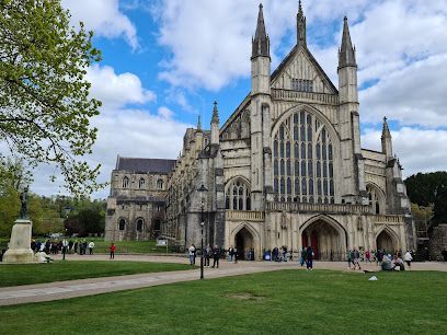 Winchester Cathedral exterior on a sunny day. Large stone building with a grassy lawn in front; people and a statue present.