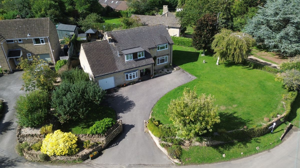 Aerial view of a two-story house with a gray roof and a green lawn, surrounded by trees and a driveway.