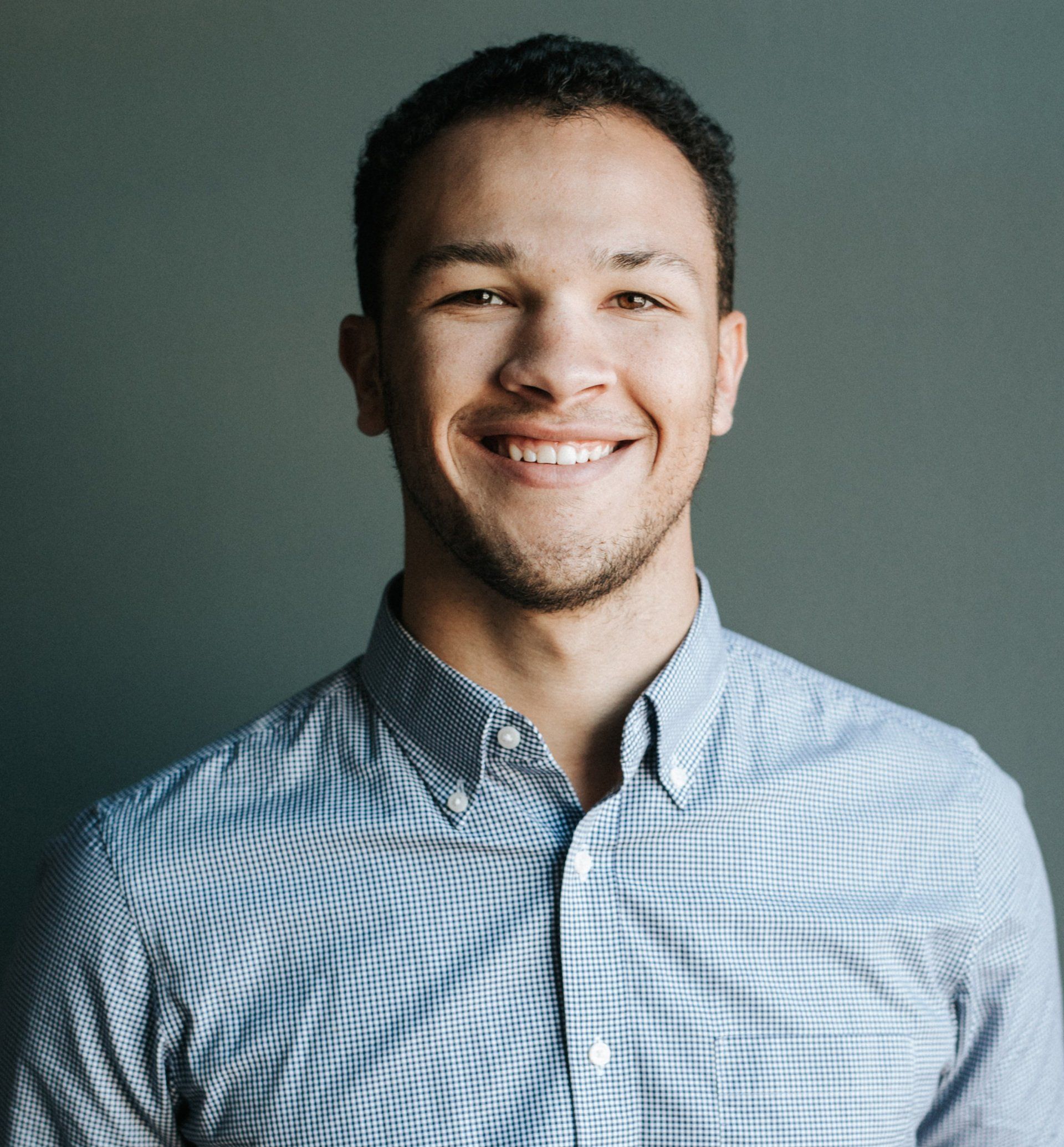 Man with dark hair smiling at the camera, wearing a blue button-down shirt against a gray backdrop.