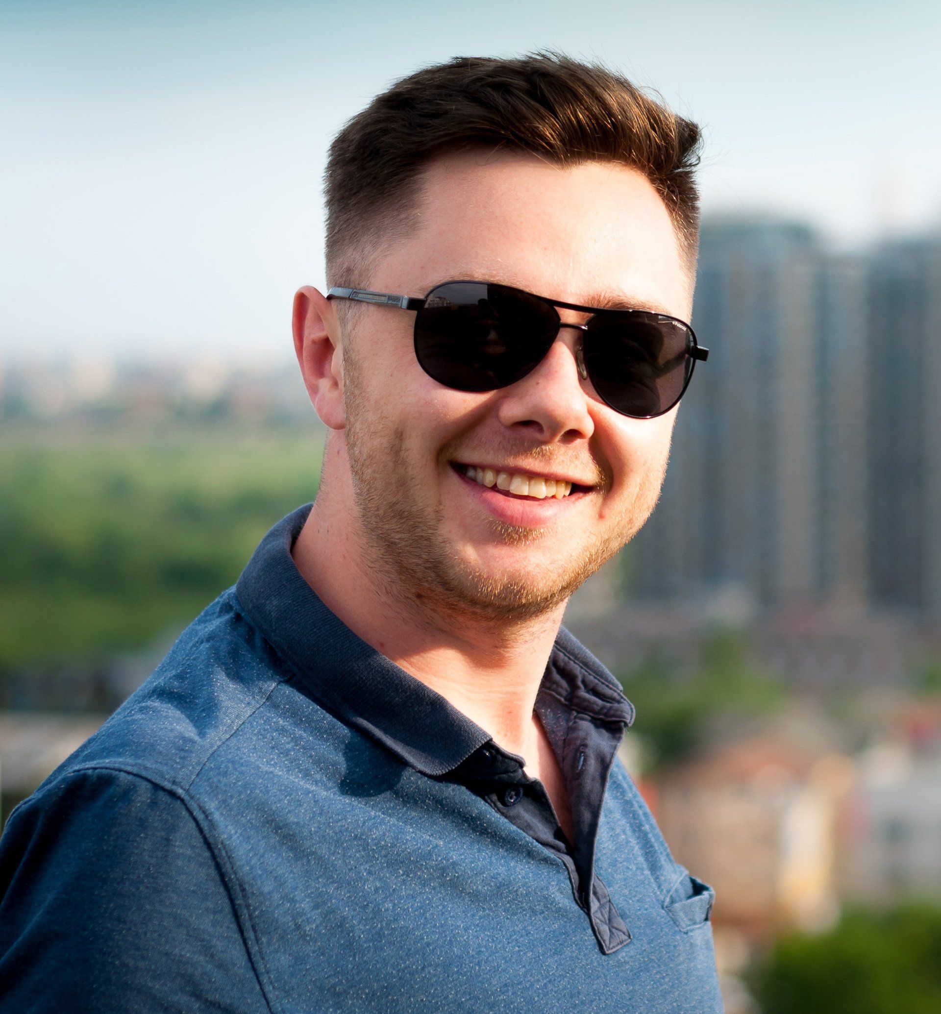 Man wearing sunglasses smiles in front of city buildings. He is wearing a blue polo.
