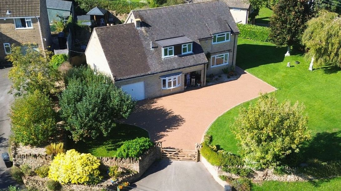 House with paved driveway and green lawn. Beige walls, dark roof. Trees and bushes around.