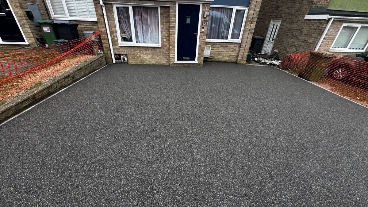 Gravel driveway in front of a brick house with windows. Red fence and a flowerbed on the sides.