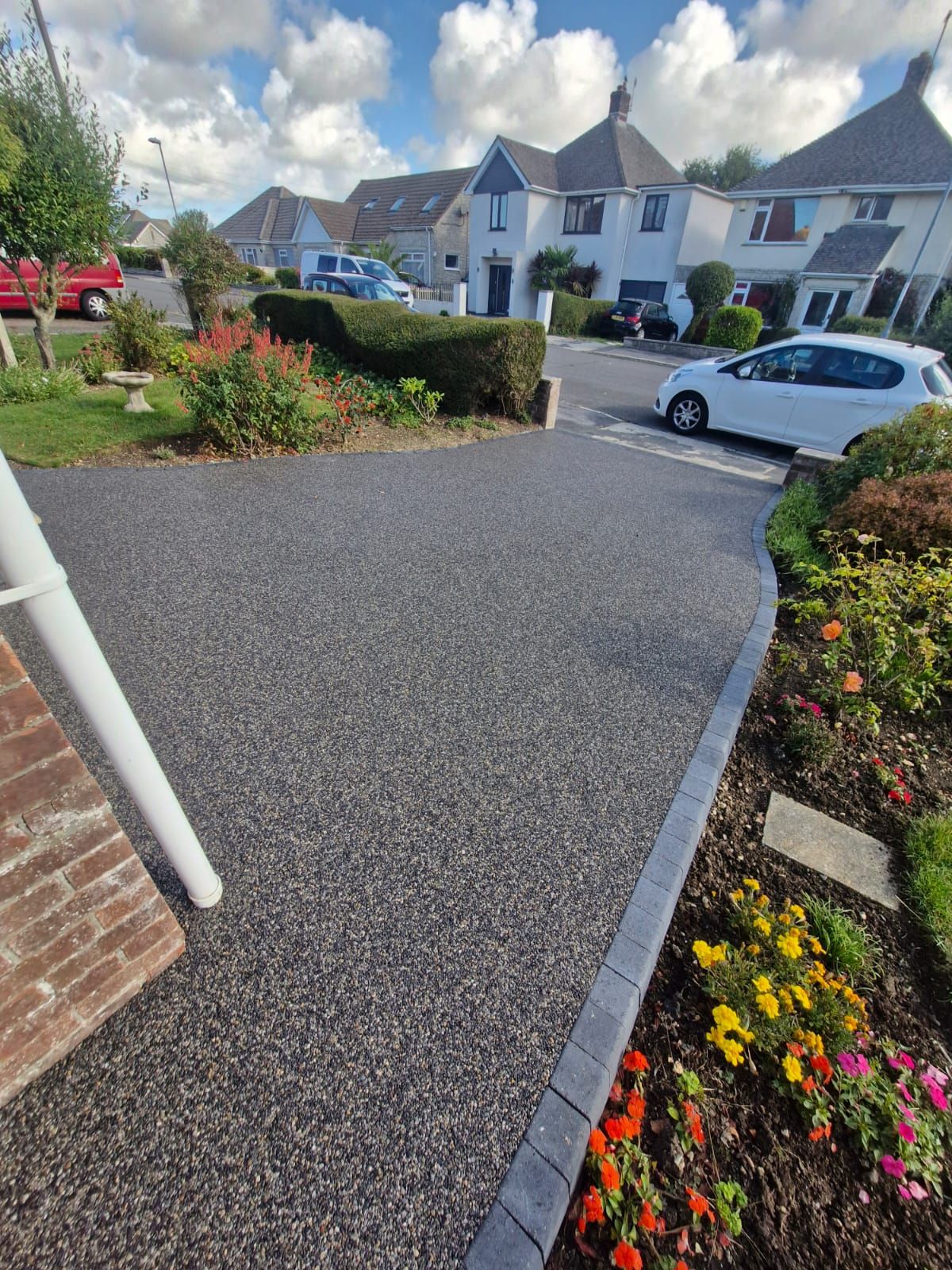 Dark gravel driveway curves past flowerbeds and houses on a sunny day.