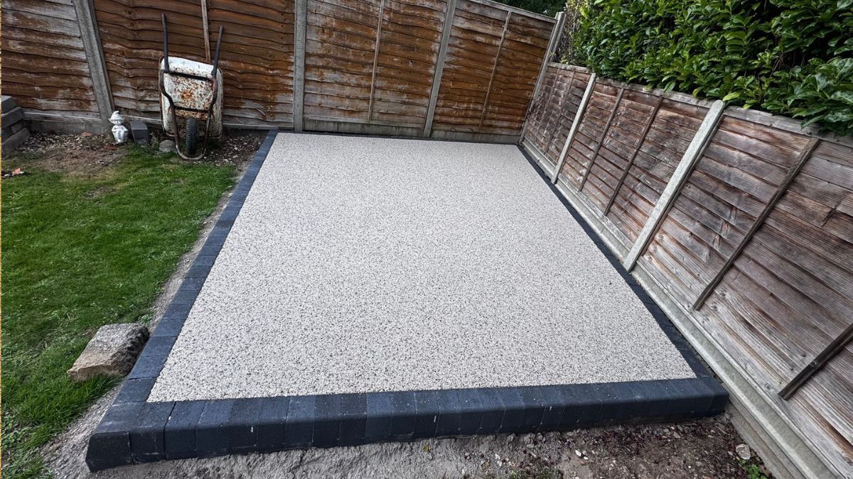A gravel patio with a dark border is bordered by a wooden fence and grass.