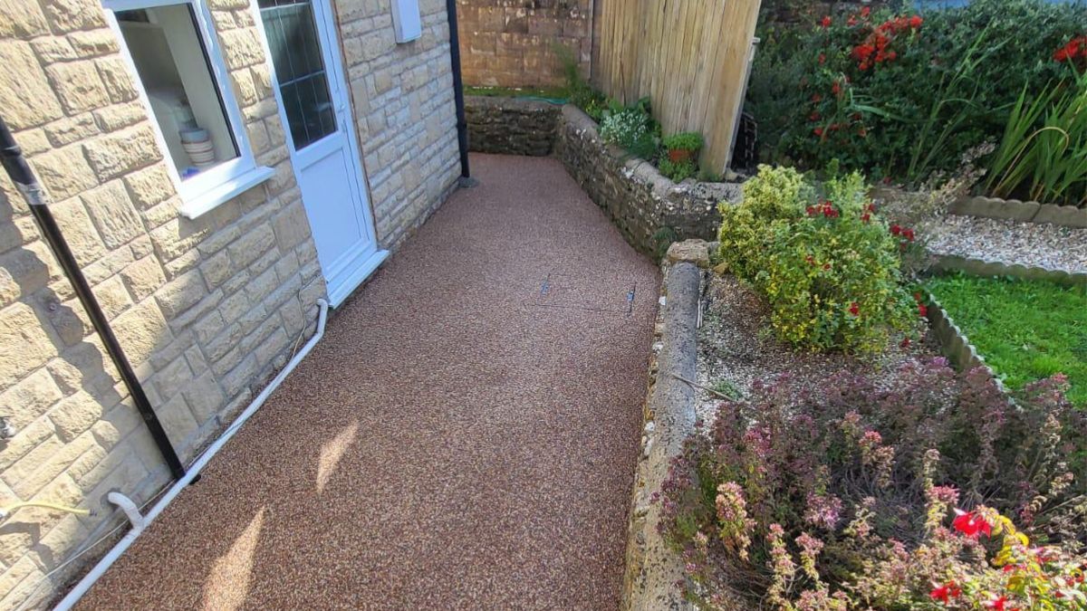 Gravel pathway beside a beige stone building and a garden bed with green shrubs and red flowers.