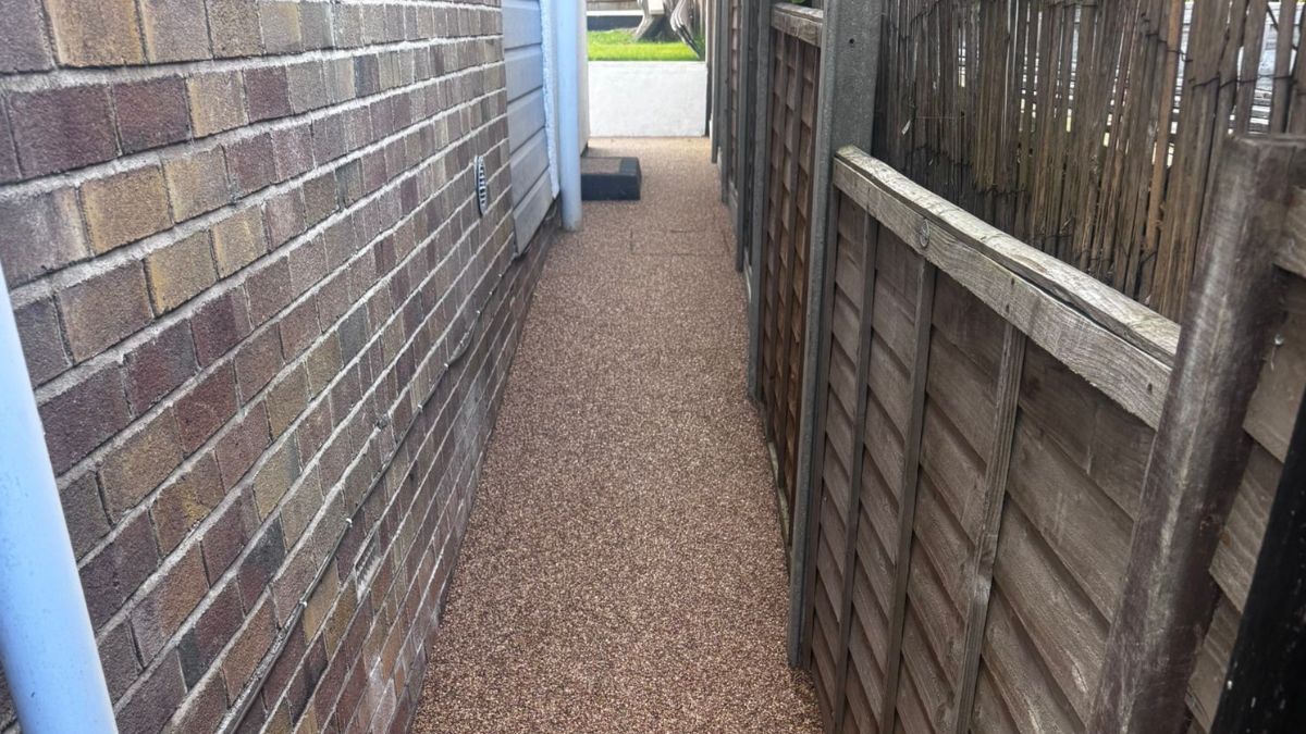Narrow pathway between a brick wall and a wooden fence, covered in small brown stones.