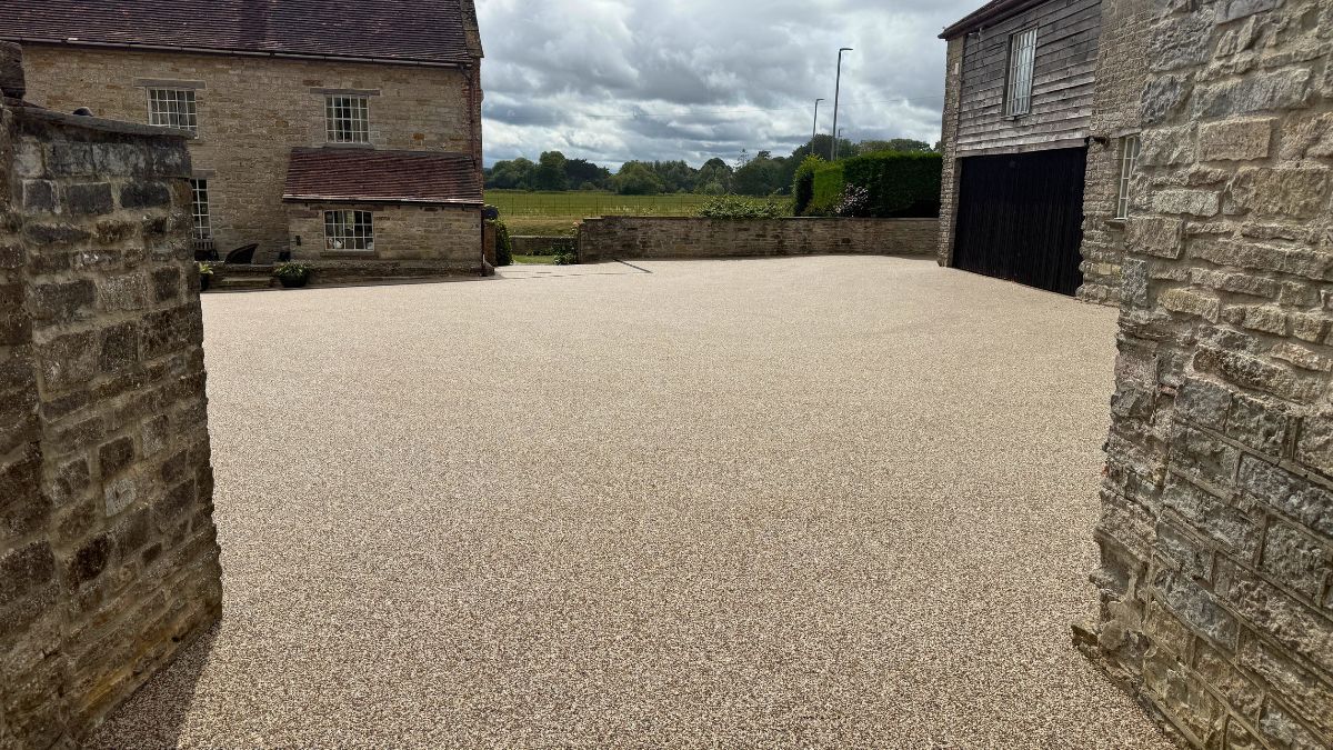 Resin courtyard between stone buildings, leading to a field under a cloudy sky.