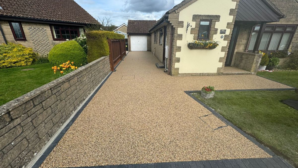 Resin bound driveway leading to a house with garage. Beige and brown hues, stone wall on the left, greenery present.