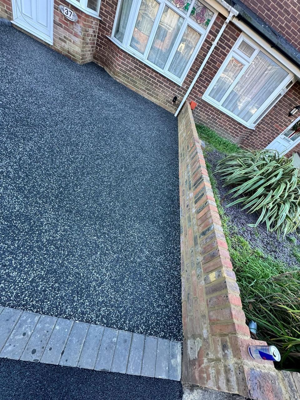 Dark driveway with brick wall next to greenery. House in background.