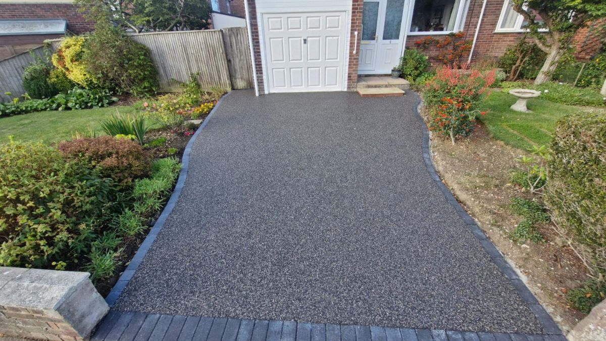 Dark grey resin driveway leading to a white garage door, flanked by gardens with greenery.
