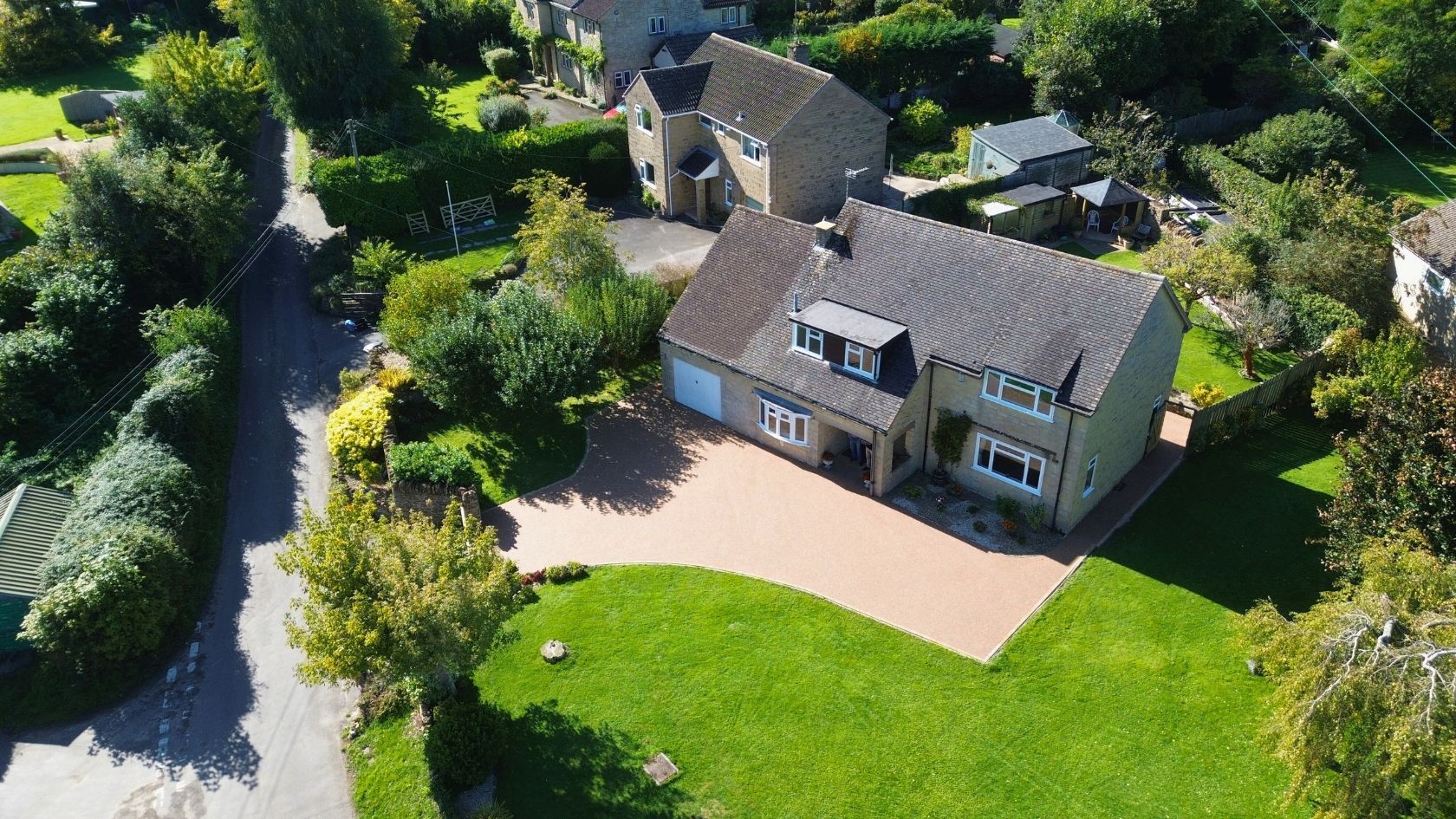 Aerial view of a two-story house with a resin bound driveway, green lawn, and surrounding trees.