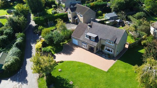Aerial view of a two-story house with a resin driveway, green lawn, and surrounding trees.