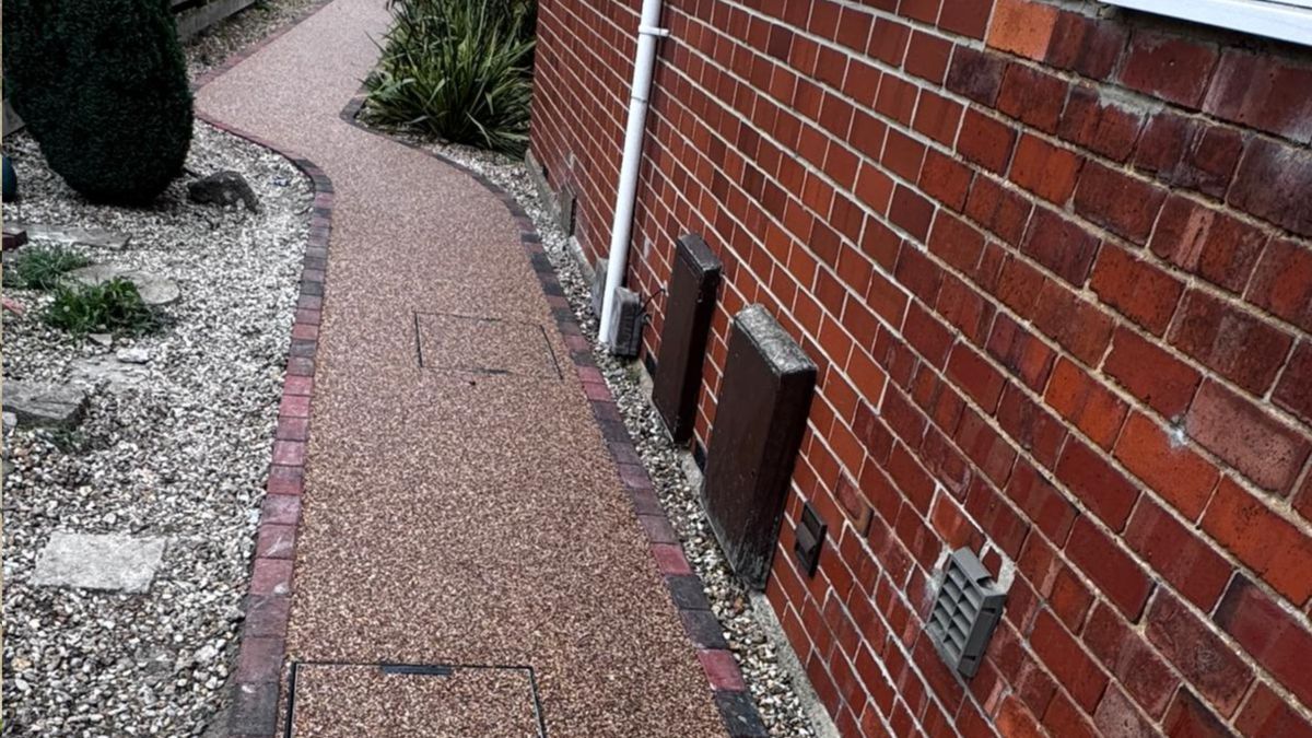 Pathway with brick edging, made of small brown stones, next to a red brick wall.