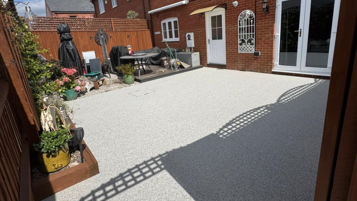 Backyard with white gravel patio, brick building, wooden fence, and shadows.