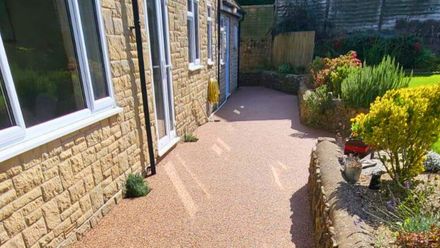 A resin paved pathway beside a brick building, edged with low walls and greenery.