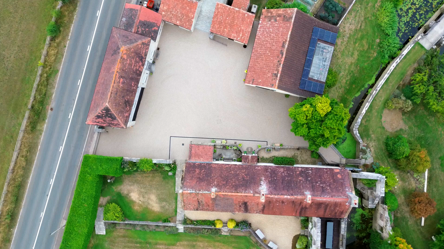 Aerial view of buildings with red tile roofs and a resin bound gravel courtyard, near a road and green space.