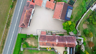 Aerial view of buildings with red tile roofs and a resin bound gravel courtyard, near a road and green space.