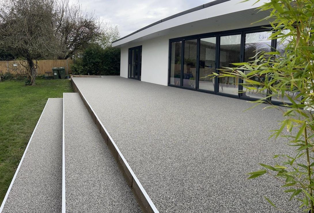 Outdoor resin patio with steps, coated in speckled gray aggregate. Glass doors of a white house in the background.