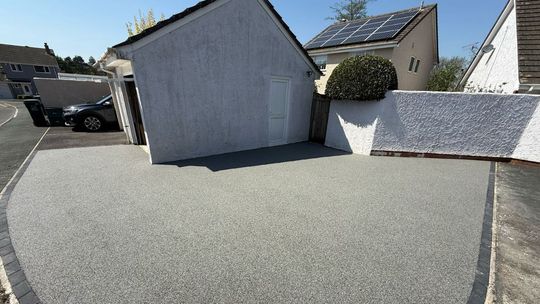 Grey resin driveway with light-colored buildings, dark car, and a wall in the background.