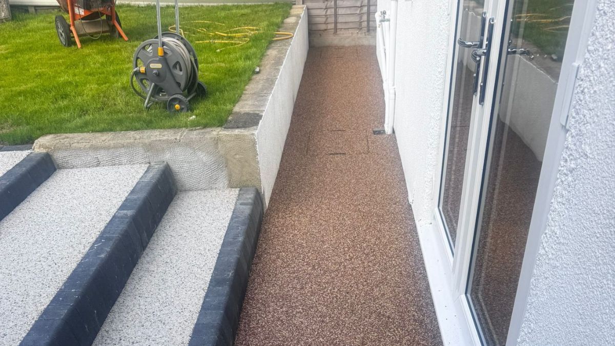 Resin-bound pathway beside a white door, next to a concrete retaining wall and grass.