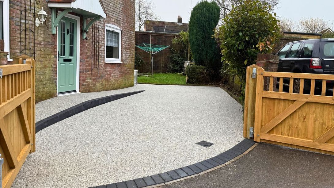 Resin front driveway with light grey colours, brick border, wooden gates, and brick house.