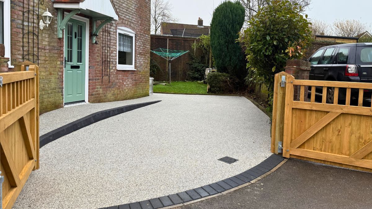 Resin front driveway with light grey colours, brick border, wooden gates, and brick house.
