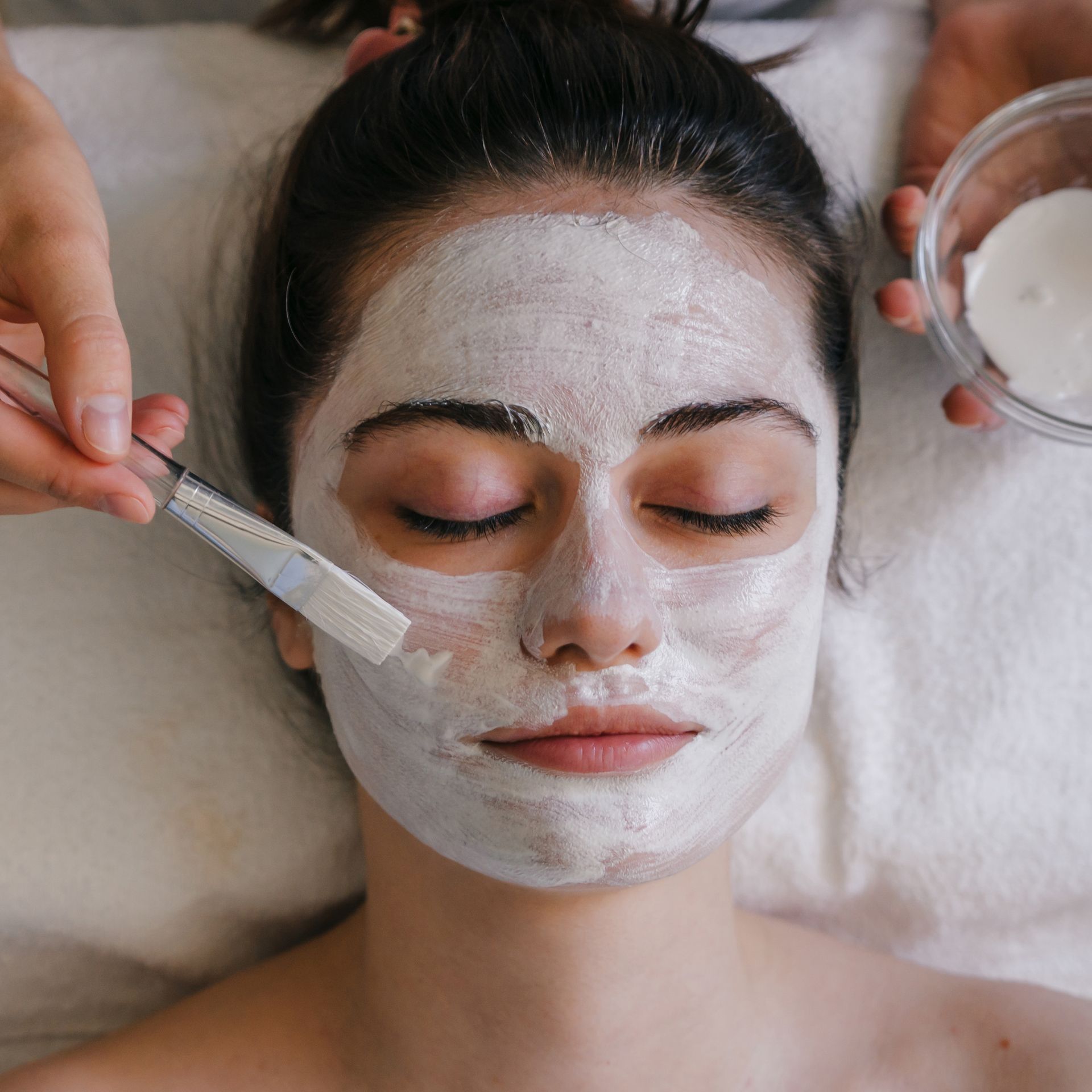 Woman getting a facial mask applied with a brush, lying down with closed eyes.