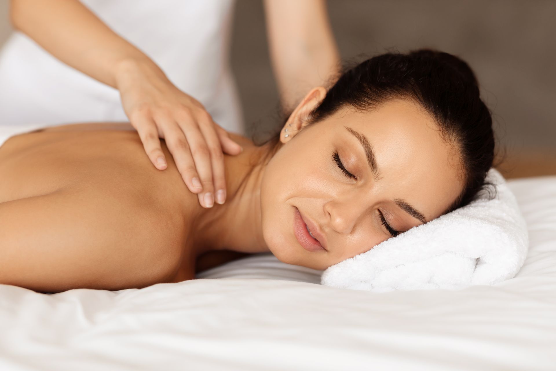 Woman receiving a back massage; therapist's hands on shoulders; lying on a white table with eyes closed, peaceful expression.