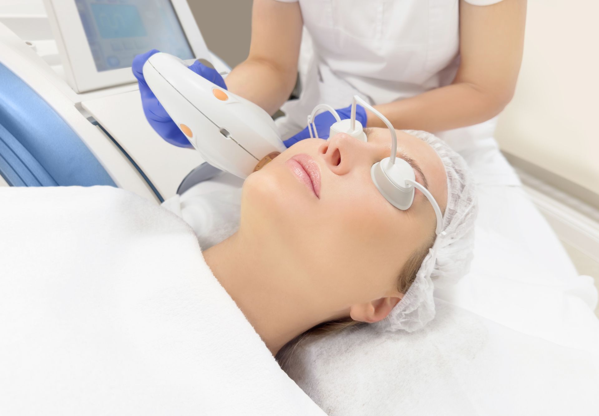 Woman in a white robe, wearing a face mask at a spa, with her hands gently on her face.