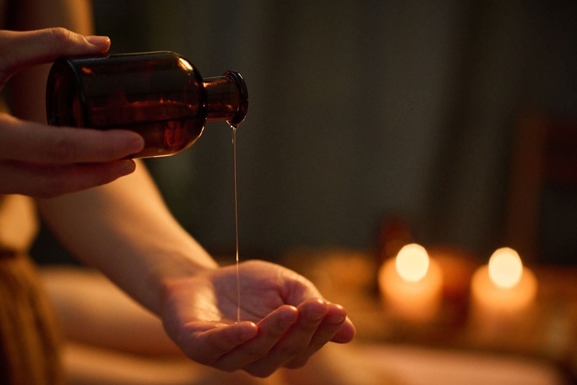 Hands pouring oil from a brown bottle into a cupped palm, lit by warm candlelight.