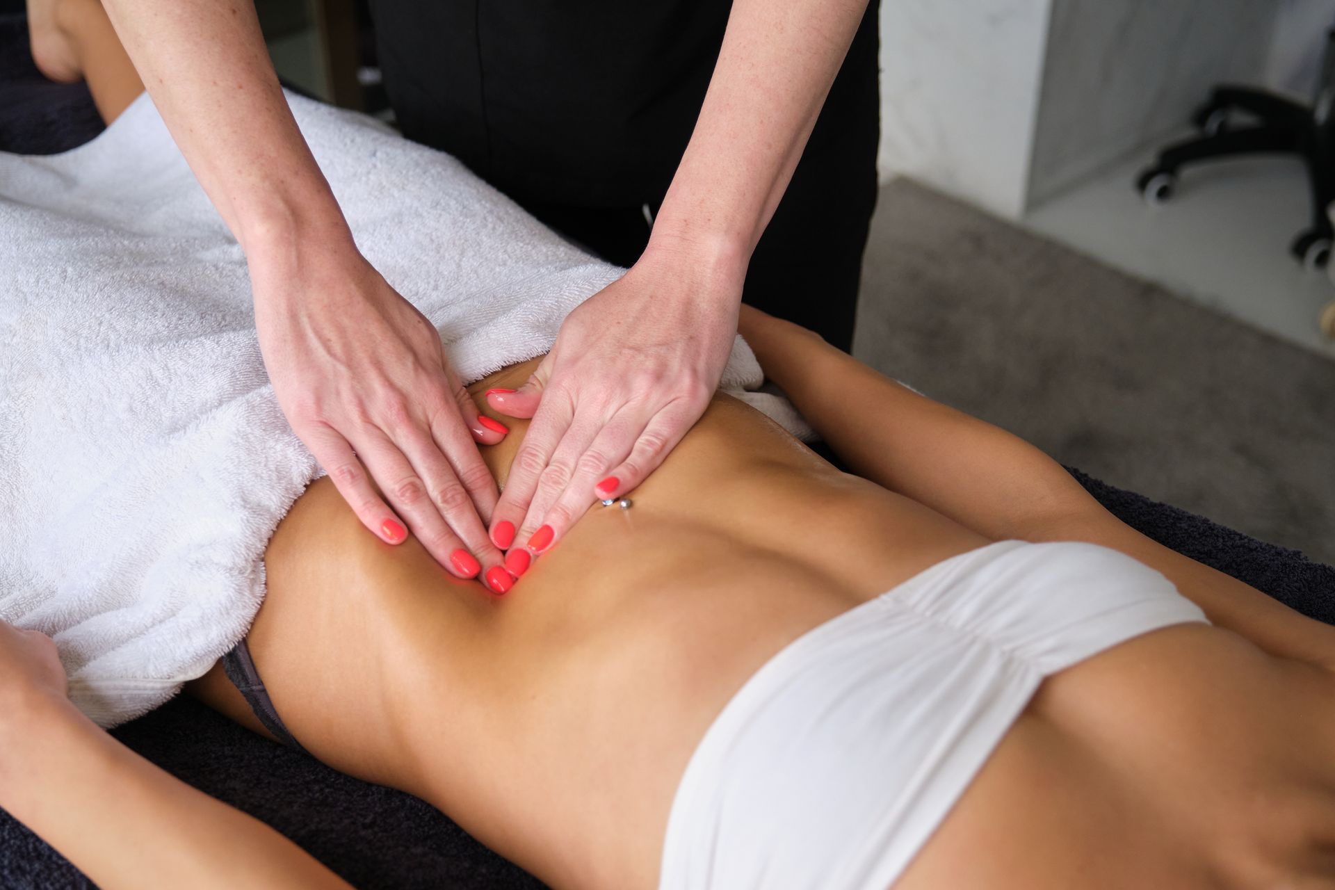 Massage therapist's hands massaging a person's abdomen, on a massage table. The person wears a white bra and is covered with a towel.