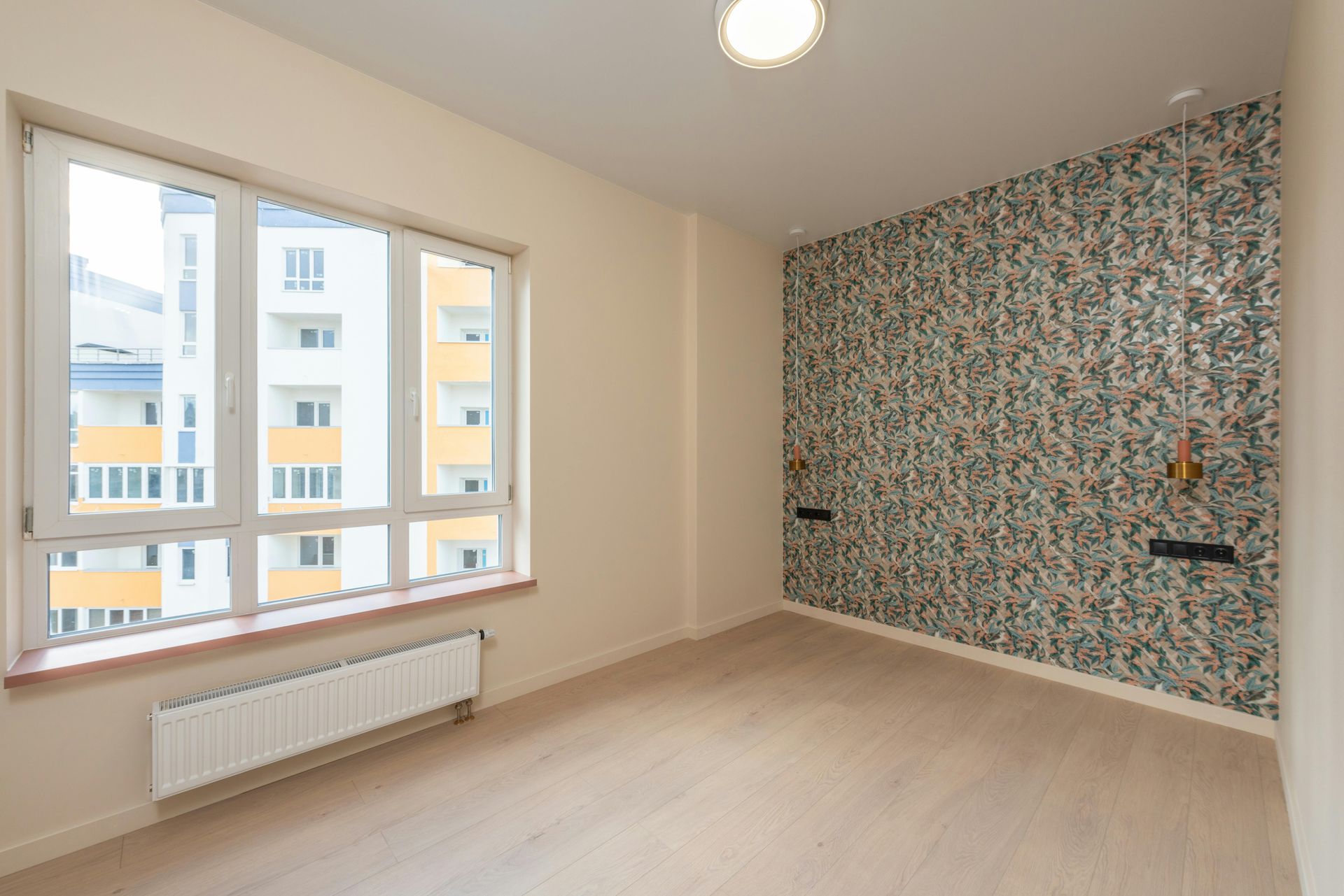 Empty room with large window, light wood floor, floral accent wall, and radiator under the window