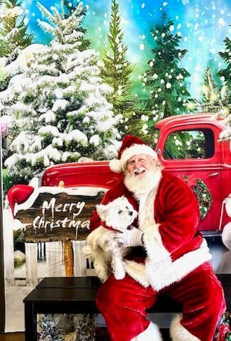 Santa Claus sitting, holding small white dog, with Christmas backdrop: red truck, snowy trees.