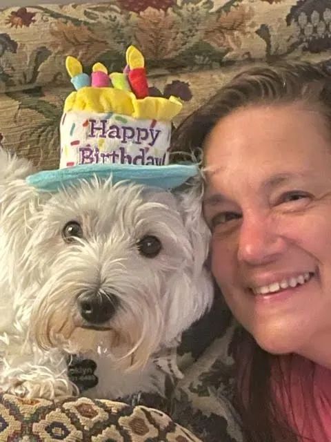 Woman smiles next to a white dog wearing a birthday cake hat.