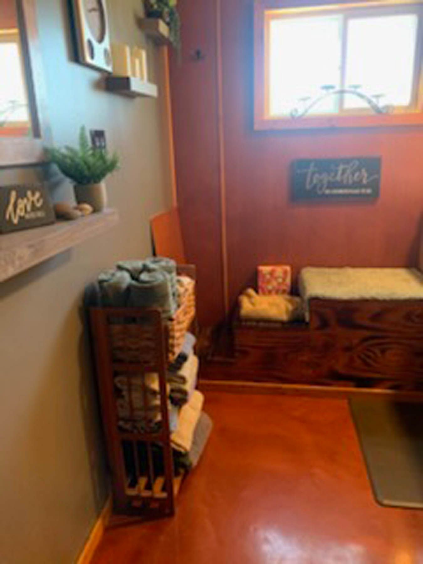 Cozy room with towels in a wooden rack, bench, and window. Warm colors: brown, gray, and red flooring.