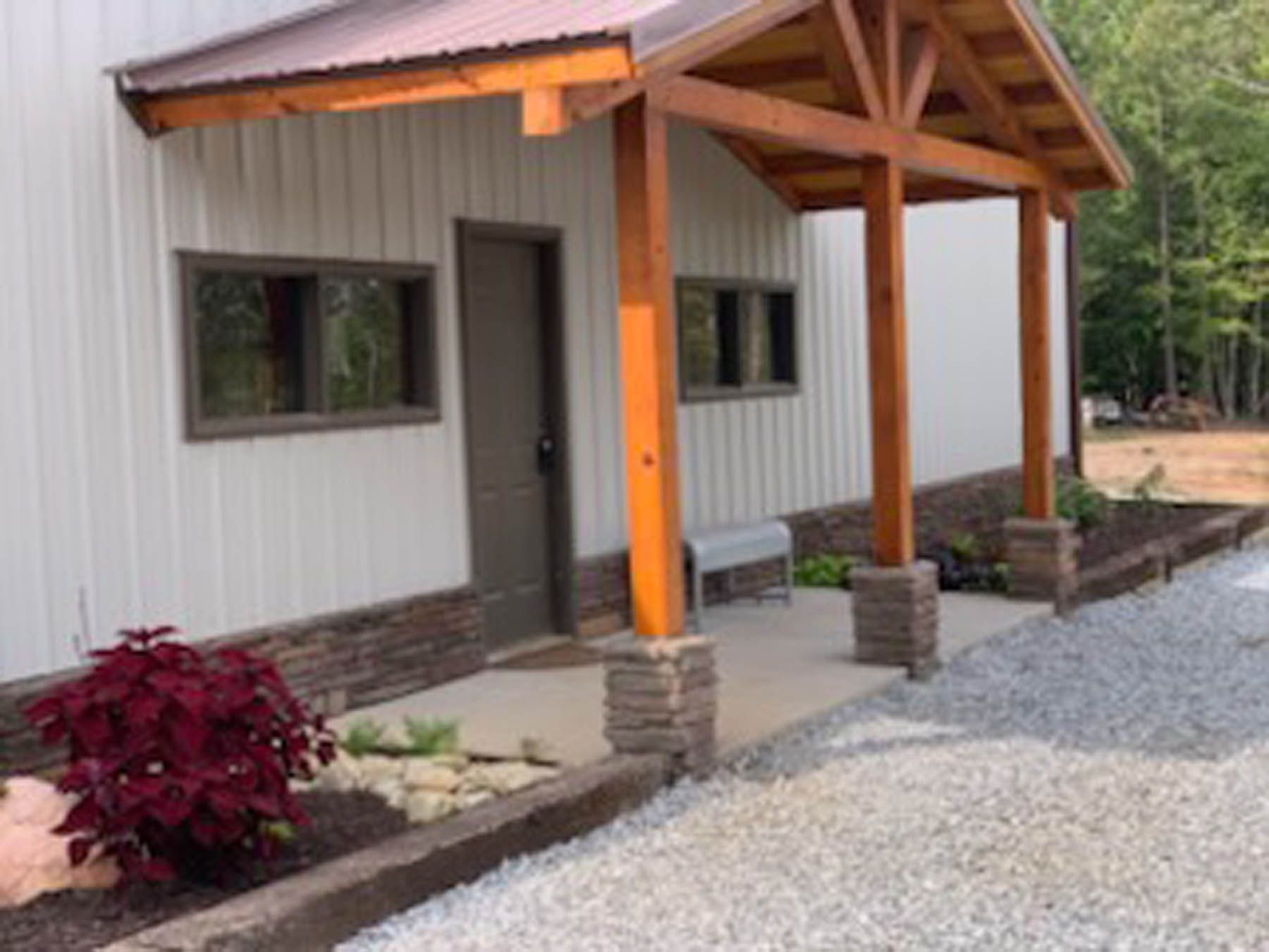 White metal building with brown wooden porch and roof, gravel driveway, and a stone planter.