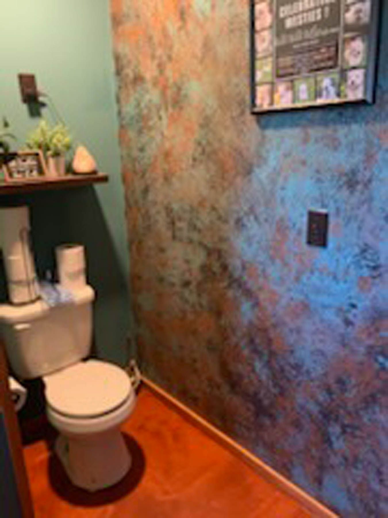 A small bathroom with a white toilet, teal and copper-colored walls, and a reddish-brown floor.